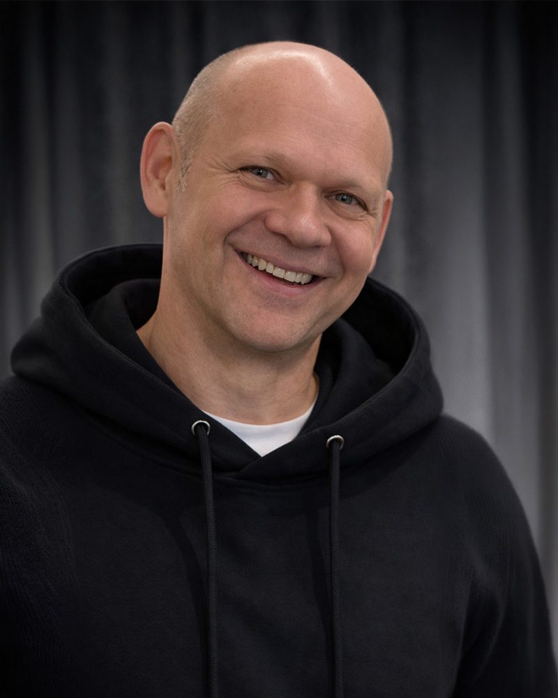 Ein nahes Porträtfoto von Carsten Grentrup, einem der Geschäftsführer unseres E-Shops für Textildruck in der Pfalz. Carsten ist kahlköpfig und blickt glücklich und lachend direkt in die Kamera. Er trägt einen schwarzen Kapuzenpullover (Hoodie) über einem weißen T-Shirt. Der Hintergrund ist ein unscharfer, dunkler Stoff.