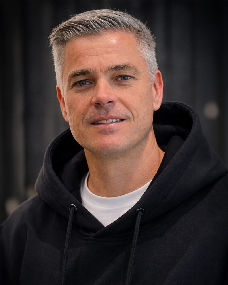 Ein nahes Porträtfoto von Christian Baist, einem der Geschäftsführer unseres E-Shops für Textildruck in der Pfalz. Christian blickt offen und lächelnd direkt in die Kamera. Er hat graumeliertes Haar und trägt einen bequemen, schwarzen Kapuzenpullover (Hoodie) über einem weißen T-Shirt. Der Hintergrund ist ein unscharfer, dunkler Stoff.