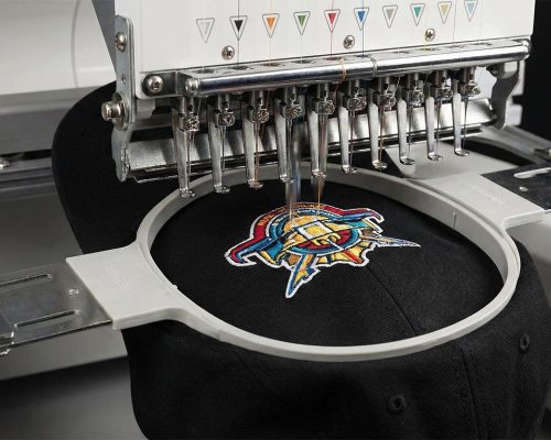 Industrielle Stickmaschine bestickt eine schwarze Cap mit einem mehrfarbigen Firmenlogo. Beispiel für hochwertige Bestickung von Caps in Bad Dürkheim.