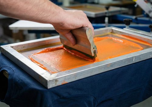 Manueller Siebdruck-Prozess: Rakel drückt leuchtend orange Farbe durch ein Sieb auf ein dunkelblaues T-Shirt – hochwertige Textilveredelung in Bad Dürkheim.