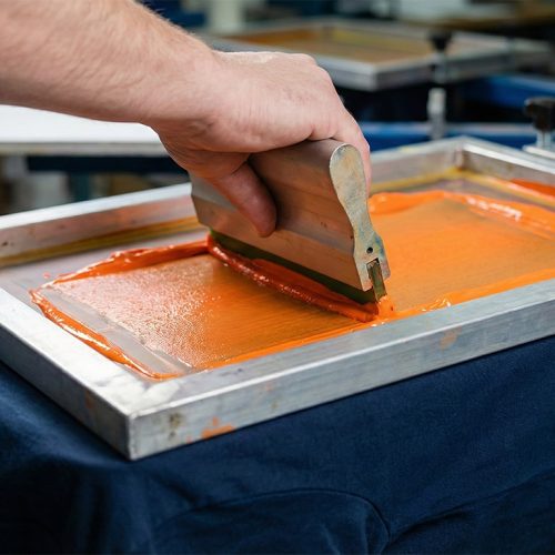 Manueller Siebdruck-Prozess: Rakel drückt leuchtend orange Farbe durch ein Sieb auf ein dunkelblaues T-Shirt – hochwertige Textilveredelung in Bad Dürkheim.