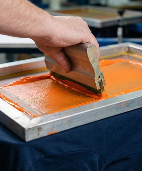 Manueller Siebdruck-Prozess: Rakel drückt leuchtend orange Farbe durch ein Sieb auf ein dunkelblaues T-Shirt – hochwertige Textilveredelung in Bad Dürkheim.