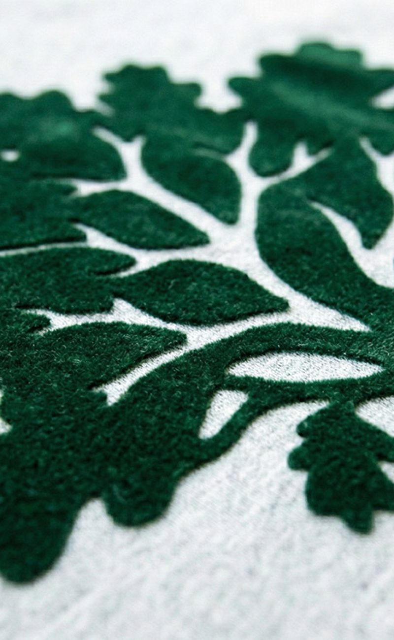 Makro-Aufnahme eines grünen Flockdrucks auf grauem Sweatshirt – deutlich erkennbare samtige Fasern und erhabene Struktur.