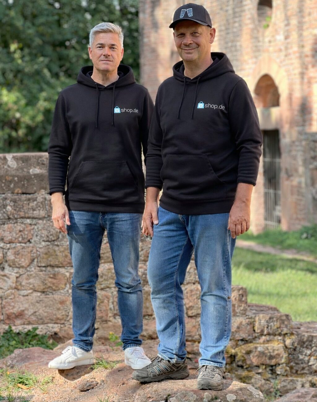 Die beiden Geschäftsführer Christian Baist (links) und Carsten Grentrup (rechts) stehen lächelnd draußen auf einer alten Steinmauer. Beide tragen identische schwarze Kapuzenpullover mit dem 'eshop.de' Logo auf der linken Brust sowie legere blaue Jeans. Carsten Grentrup trägt zusätzlich eine schwarze Basecap.