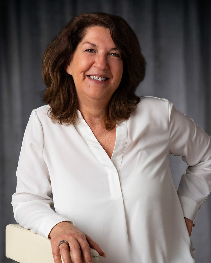 Ein freundliches Porträtfoto von Jutta Großheinrich, unserer Textil-Fachberaterin im E-Shop für Textildruck in der Pfalz. Sie lächelt offen und sympathisch in die Kamera, trägt eine elegante weiße Bluse und stützt sich entspannt auf eine helle Lehne. Im Hintergrund ist ein weicher, grauer Vorhang zu sehen.