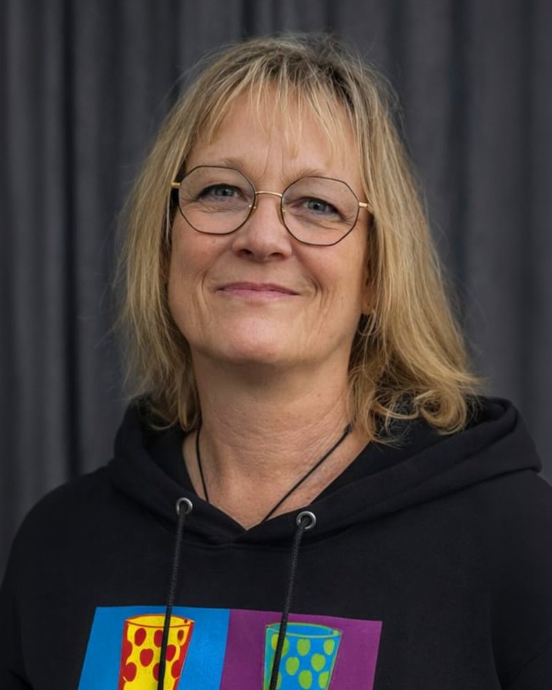 Ein hochauflösendes, professionelles Porträtfoto von Michaela Boller, einer Mitarbeiterin im E-Shop für Textildruck. Sie hat hellblondes, schulterlanges Haar und trägt eine markante achteckige Metallbrille. Sie lächelt direkt und herzlich in die Kamera. Sie trägt einen schwarzen Kapuzenpullover mit Kordelzug. Auf der Brust ist ein detaillierter Grafikdruck zu sehen: zwei Pop-Art-ähnliche, punktierte Quadrate. Das linke Quadrat ist rot-rosa mit einem punktierten Kelch-Motiv; das rechte Quadrat ist lila-purpur mit einem punktierten Kelch-Motiv. Im Hintergrund ist ein dunkler, vertikal plissierter Vorhang zu sehen.
