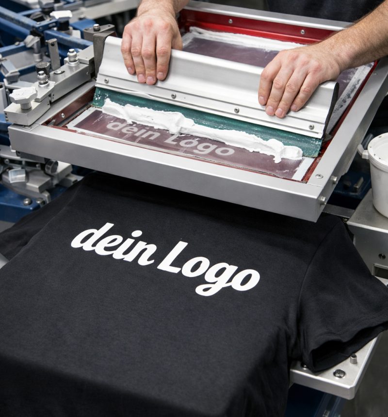 Blick in die Produktion: Manueller Siebdruck auf ein schwarzes T-Shirt. Ein Mitarbeiter zieht weiße Farbe mit dem Rakel über das Sieb für ein perfektes Druckergebnis.